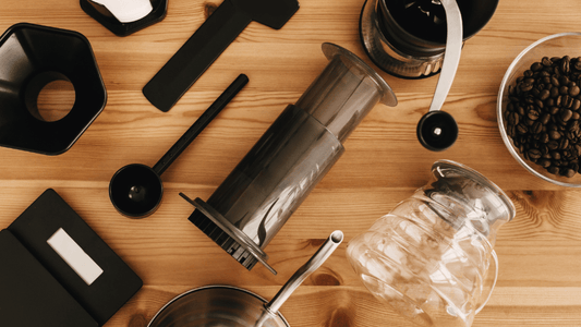 Auf einem Tisch liegt eine AeroPress samt Zubehör, einer Kaffeewaage, Kaffeebohnen und einer Karaffe.