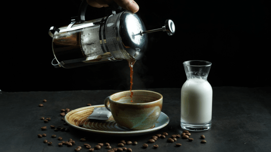 Jemand gießt frischen Kaffee aus der French Press in eine Tasse, daneben steht ein Glas mit Zucker